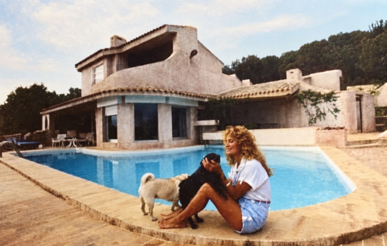 Dalida sentada junto a la piscina con sus dos perros, con la casa detrás